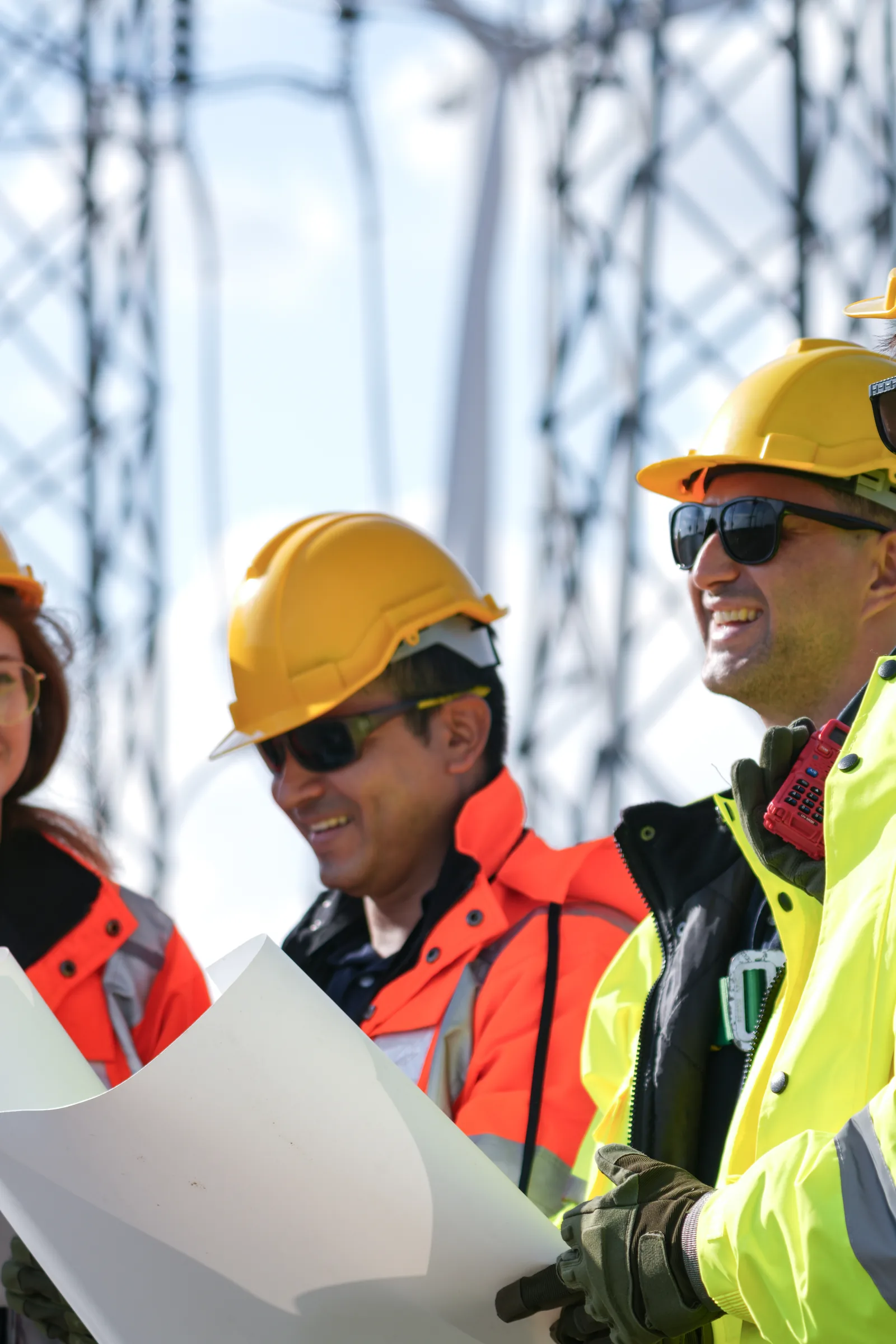 Three engineers in high-visibility clothing and hard hats reviewing a site drawing beneath a high-voltage transmission pylon, with a wind turbine behind them