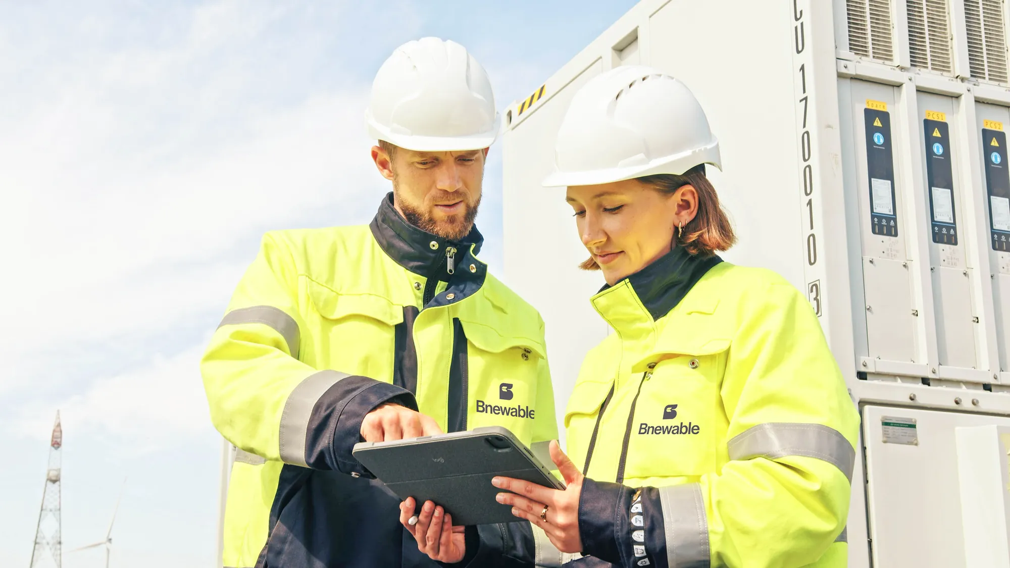 Two Bnewable engineers in high-visibility jackets reviewing battery storage data on a tablet beside a battery container