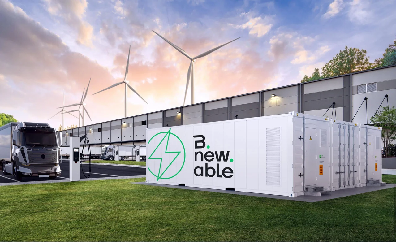 A white Bnewable battery container in a grass field, with an electric truck at a charging point, wind turbines and an industrial building in the background
