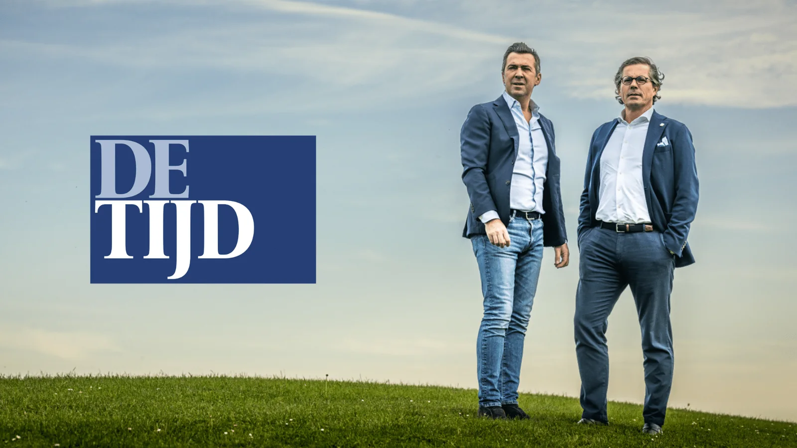Two Bnewable colleagues standing outdoors on a grassy embankment under a cloudy sky, with the De Tijd logo
