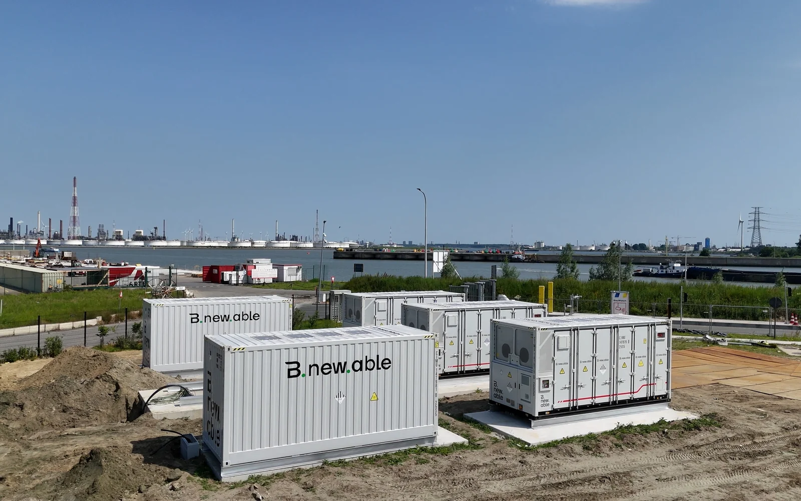 White Bnewable battery containers on a gravel construction site in the Port of Antwerp, with refinery installations and a high-voltage pylon in the background