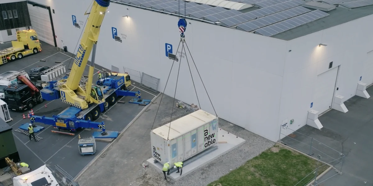 Ein Kran hebt einen Bnewable-Batteriecontainer neben einer Industriehalle mit Solardach auf sein Fundament