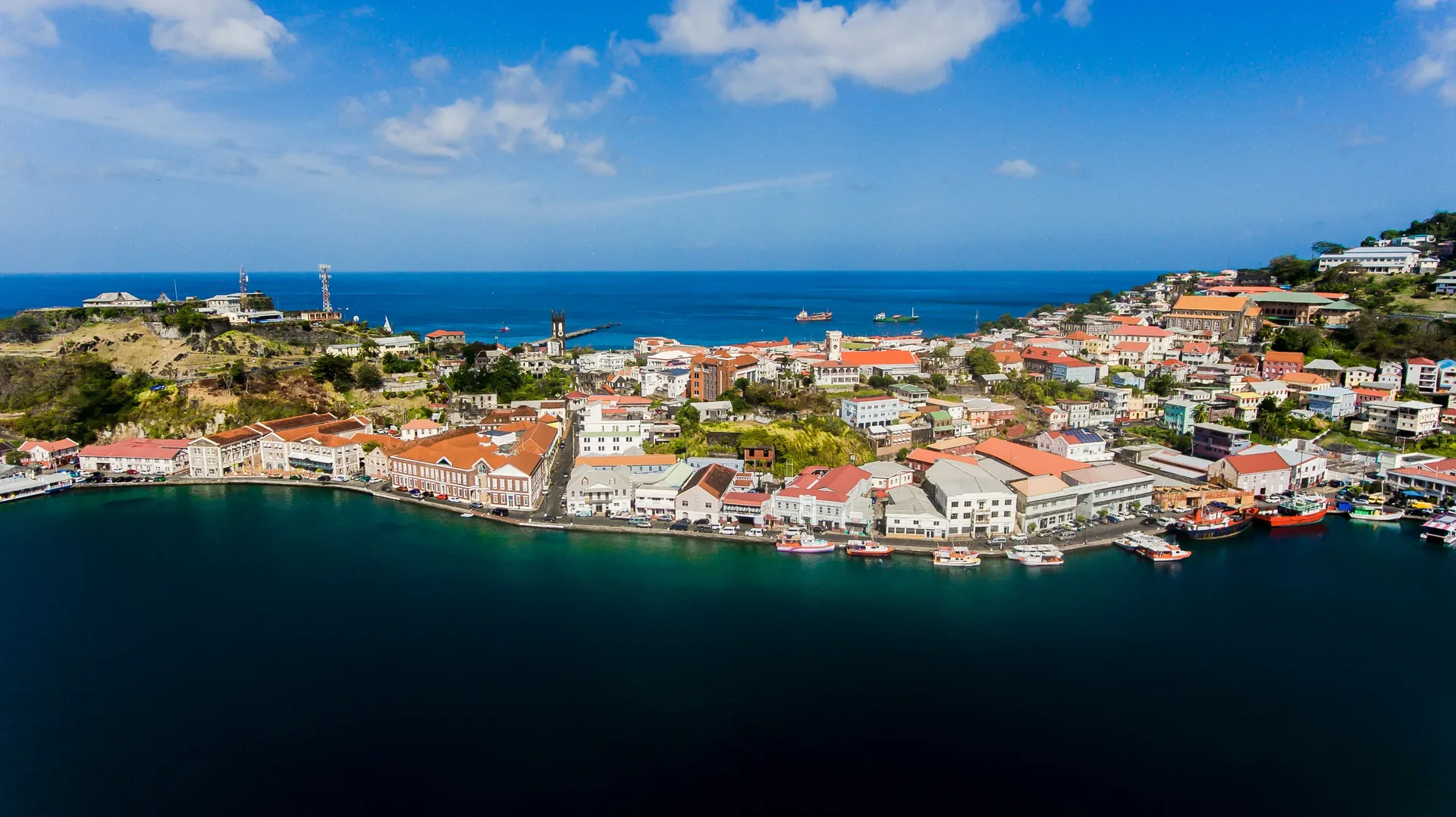 Aerial view of a coastal town with red-roofed buildings, a dock, and blue ocean under a partly cloudy sky.