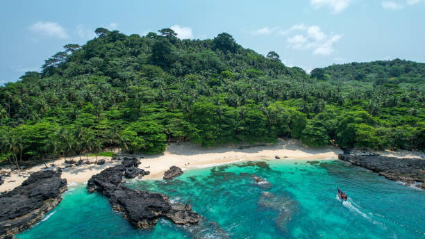 Tropical beach with clear turquoise water, black rocks, sandy shore, and dense green forested hills.