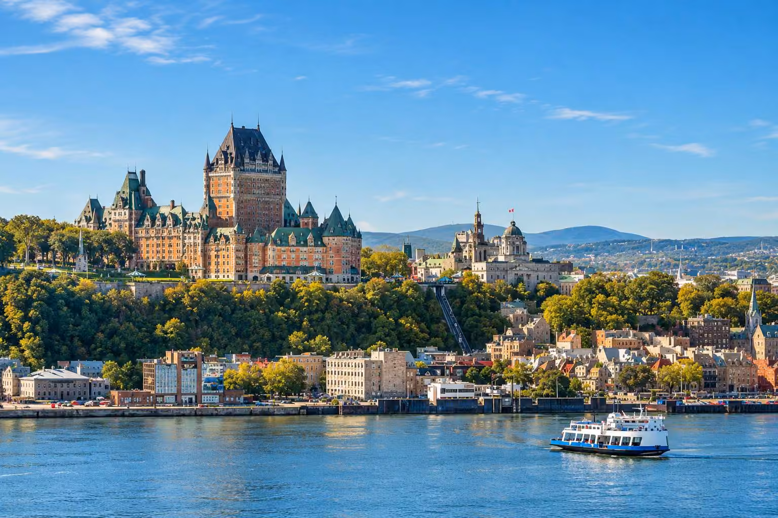 View of Quebec City with Château Frontenac castle atop a green hill, historic buildings below, and a boat sailing on the river under a clear blue sky.