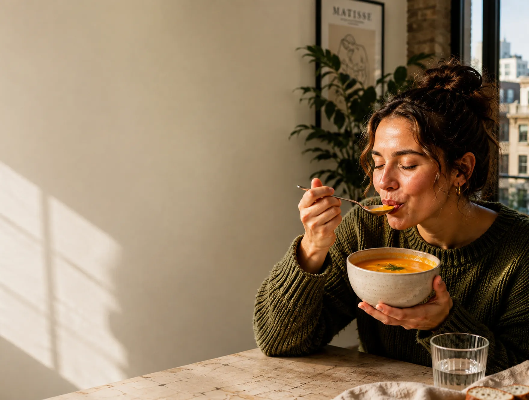Vrouw in groene trui geniet van een lepel soep terwijl ze een kom vasthoudt aan een tafel.