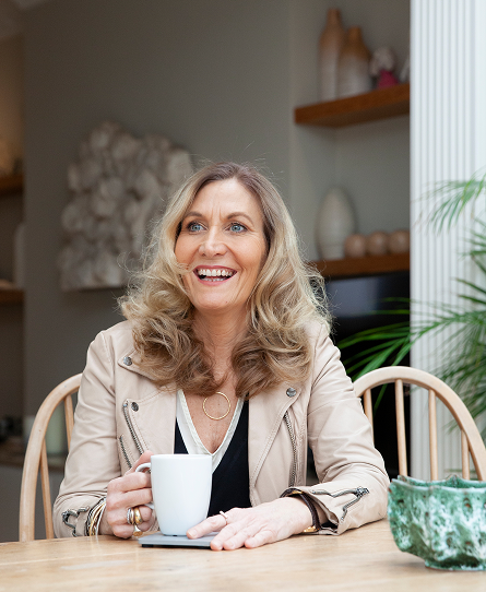 Harriet Young at a table with a coffee in hand. Smiling at someone off camera.