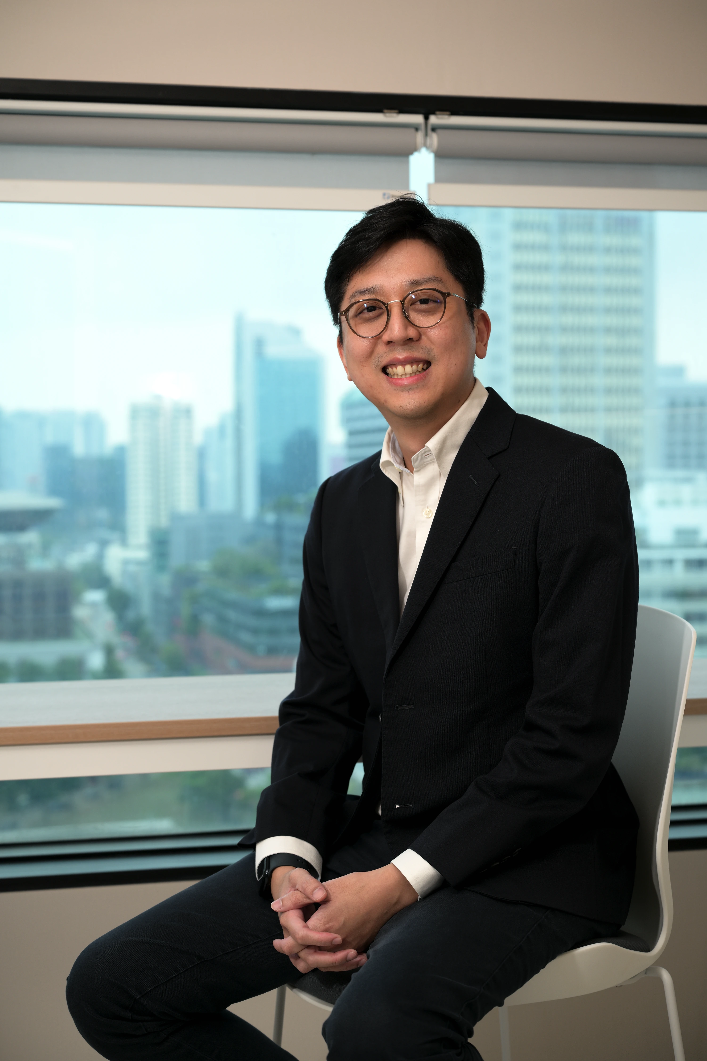 Smiling man with glasses in black suit and white shirt sitting on a chair by a window with city view.