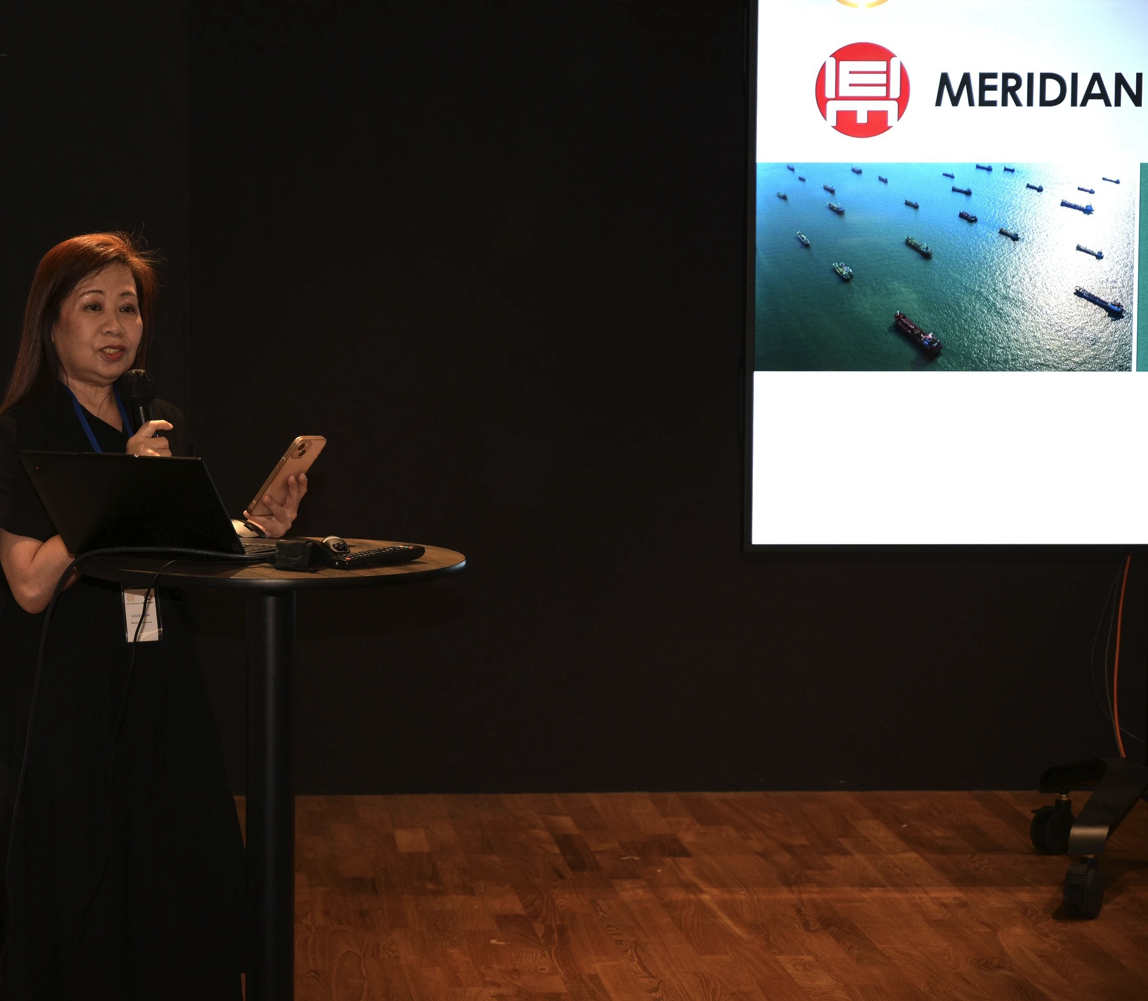 Woman holding microphone and phone, standing at a round table with laptop, speaking in front of a presentation screen.