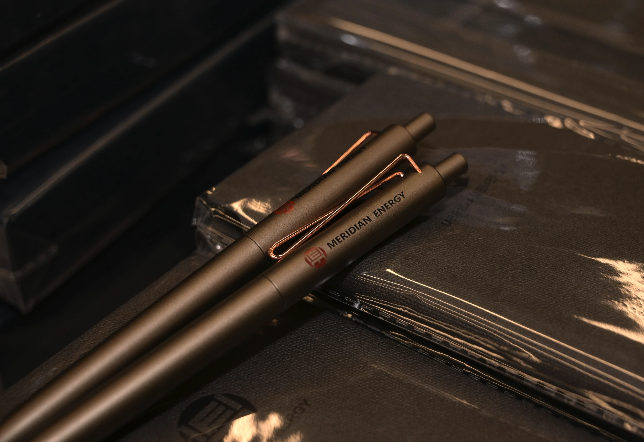 Two brown pens with 'MERIDIAN ENERGY' on them lying on stacked plastic-wrapped notebooks.