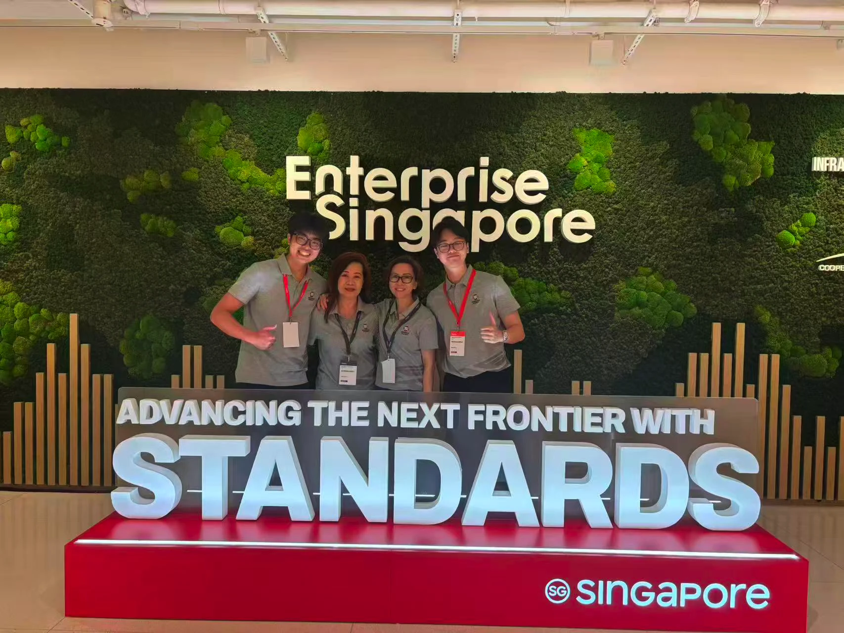 Four people in gray shirts with badges standing behind a lit sign saying advancing next frontier with standards.