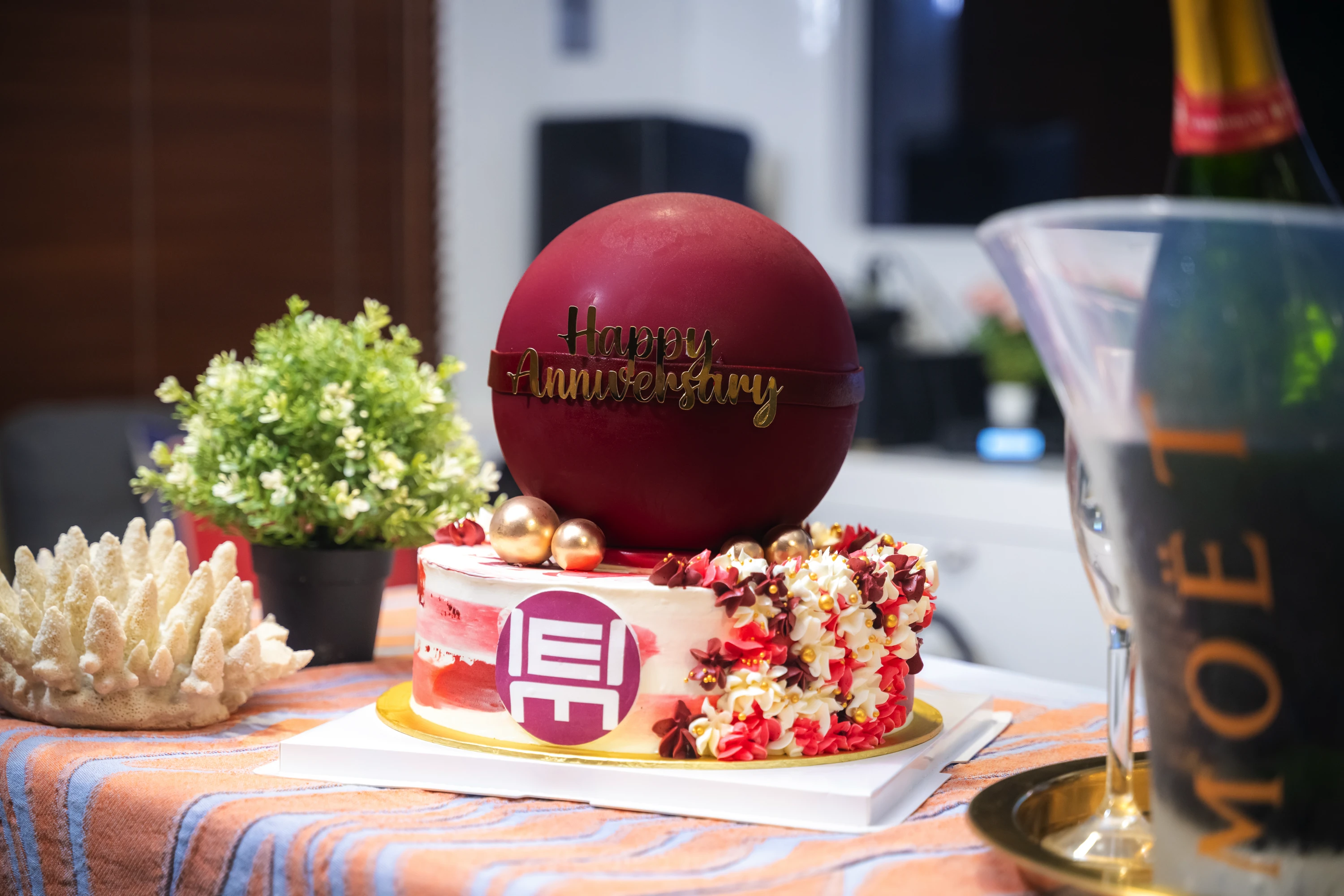 Anniversary cake with a large red chocolate ball, flowers, gold spheres, and Happy Anniversary sign.