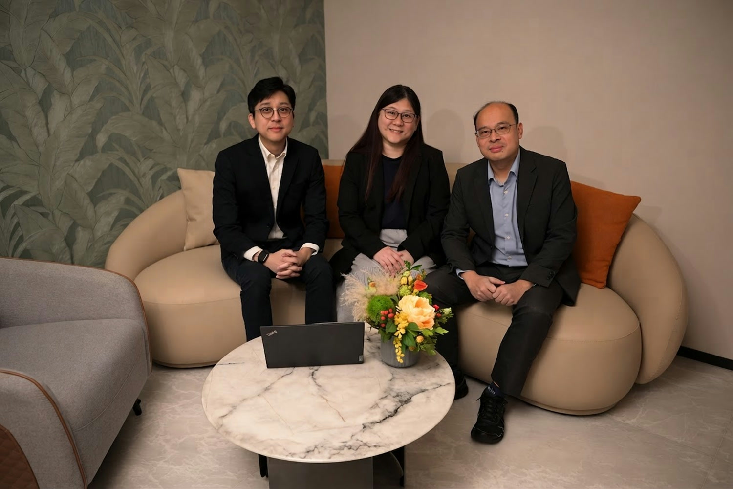 Three professionals sitting on a beige sofa with a laptop and flower vase on a round marble table.