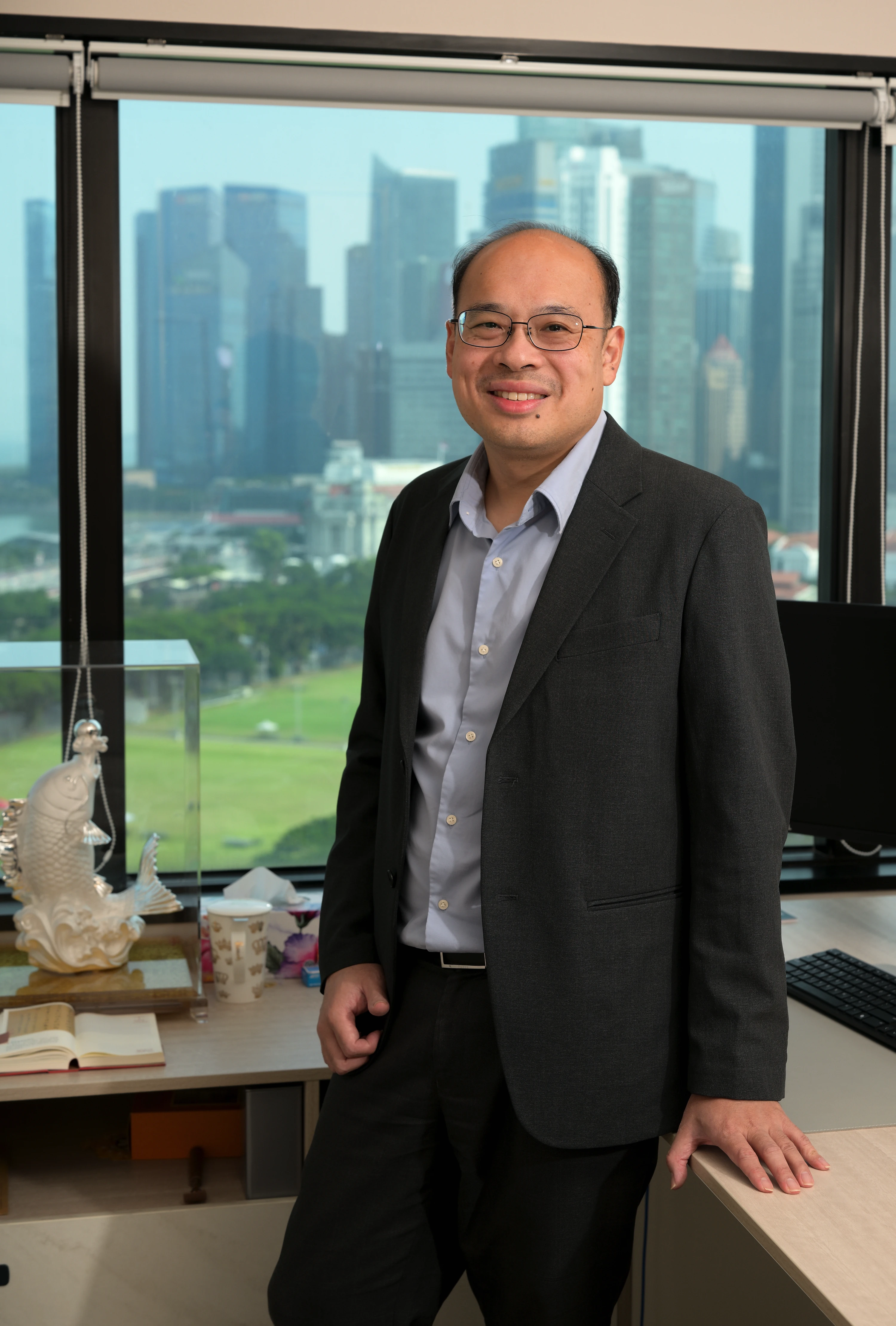Man in glasses and dark suit stands smiling by office desk with city skyline in background.