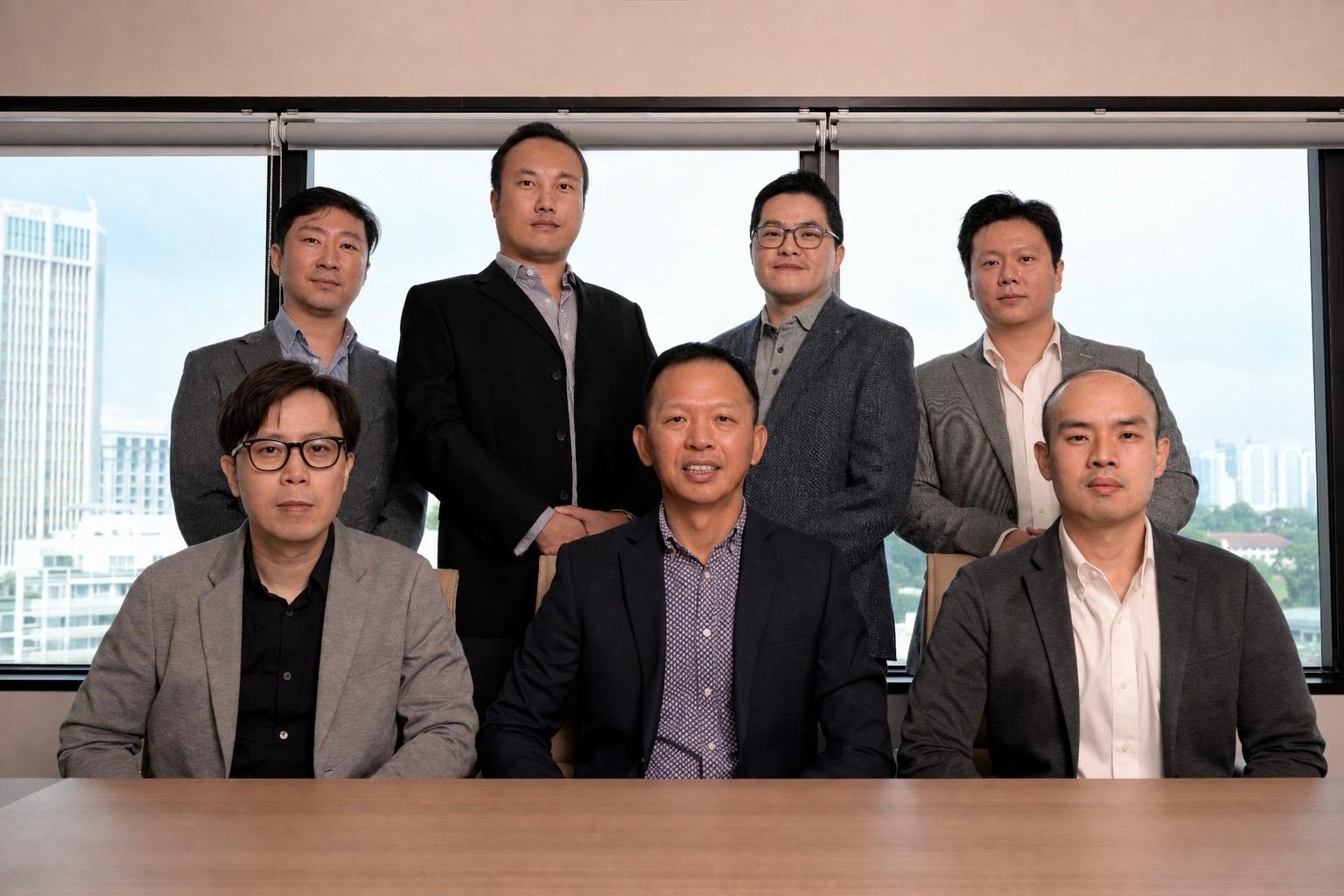 Group of seven Asian men in business attire posing in front of office window with city view.