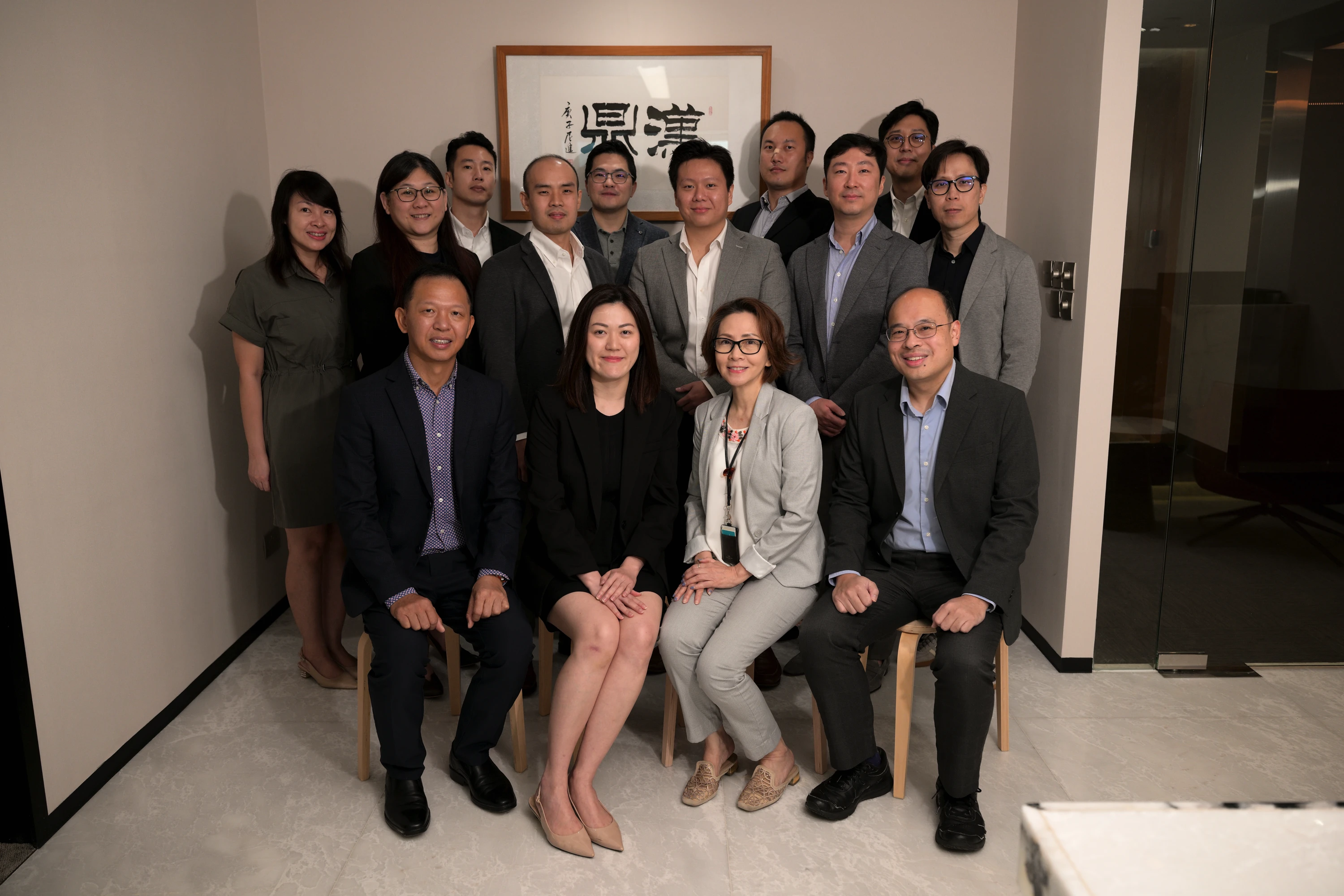 Group of thirteen professionally dressed people posing in an office with framed calligraphy on the wall.