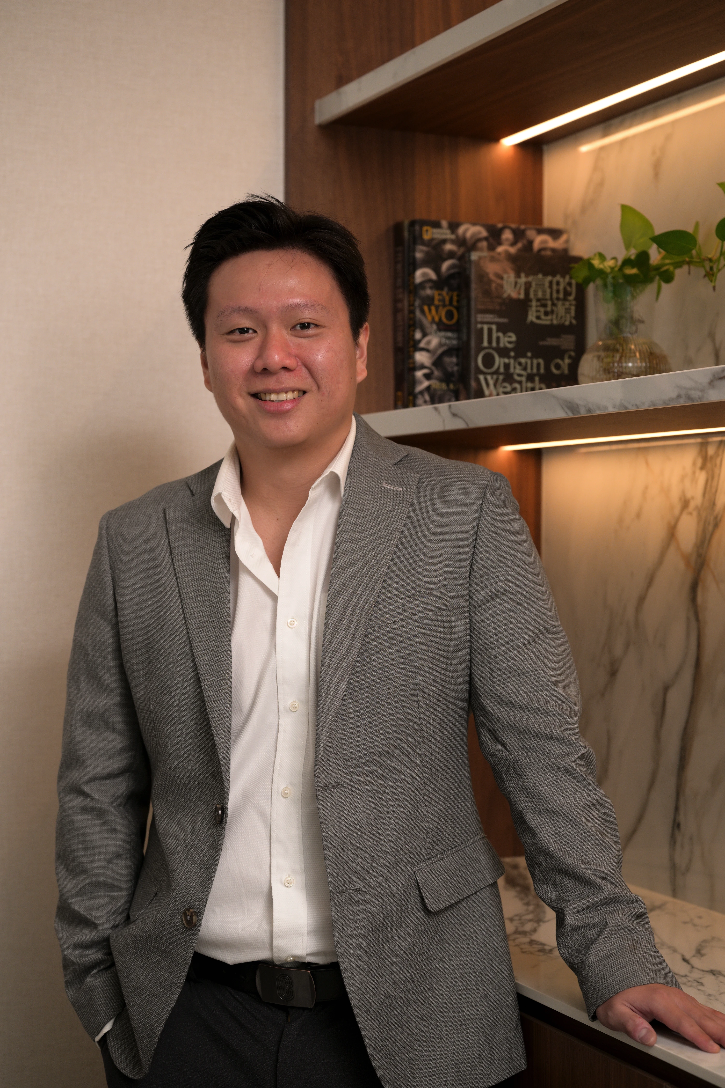 Smiling man in gray blazer and white shirt leaning on a marble countertop with bookshelves in background.