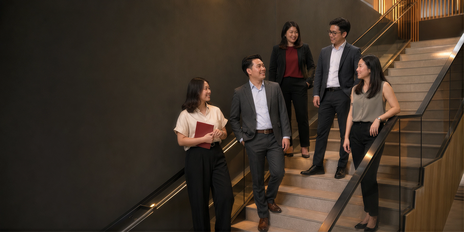 Five professionally dressed people standing and chatting on stairs with dark wall and glass railing.