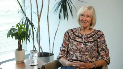 Smiling older woman sitting indoors with patterned blouse and plants in the background.