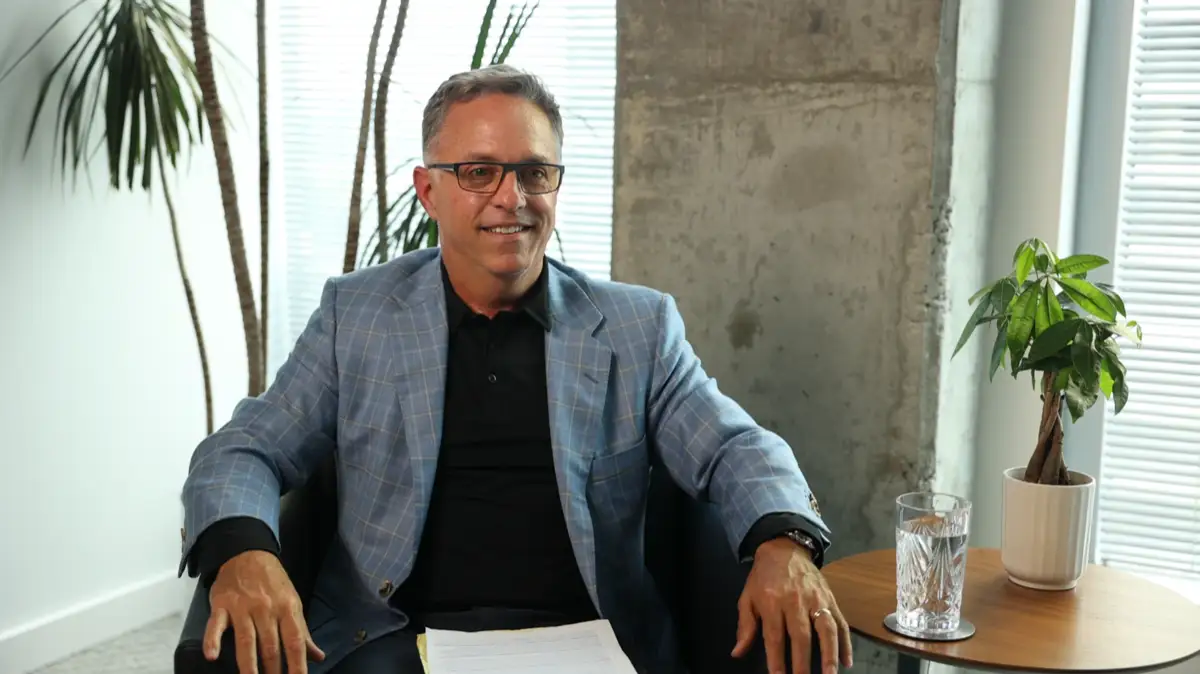 Man in glasses and checkered blazer sitting and smiling in a modern room with plants and a glass of water.