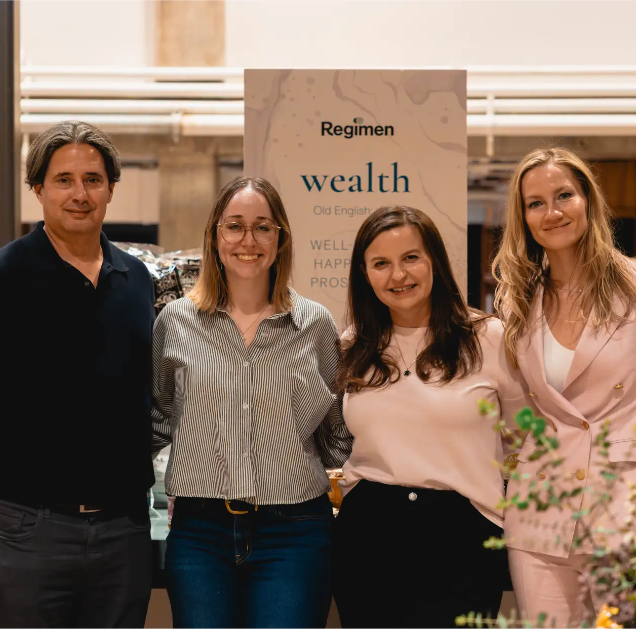 Four smiling adults standing together in front of a 'Regimen wealth' sign indoors.