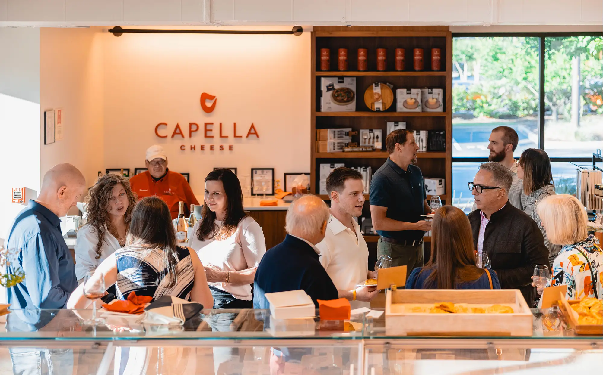 Group of people talking inside Capella Cheese shop with products and glass display counters visible.