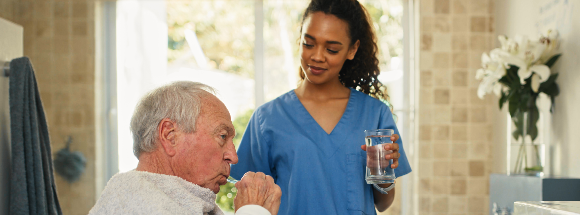Elderly man wearing a towel around his shoulders brushing teeth while a healthcare worker in blue scrubs holds another glass of water and looks on with a gentle smile.