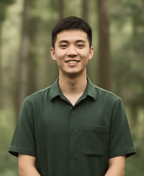 Smiling young man wearing a dark green collared shirt standing outdoors with blurred trees in the background.