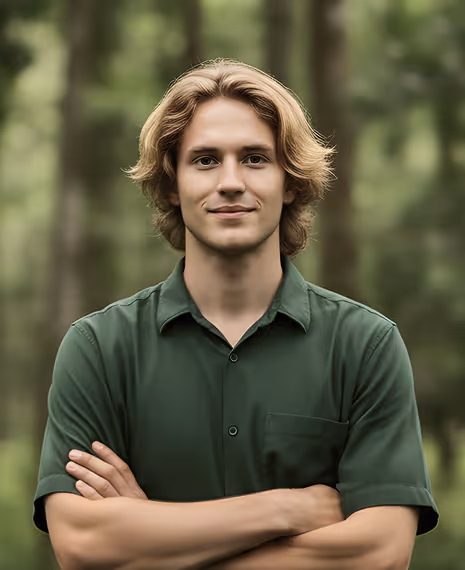 Young man with blonde hair wearing a green shirt, standing outdoors with arms crossed and trees in the background.