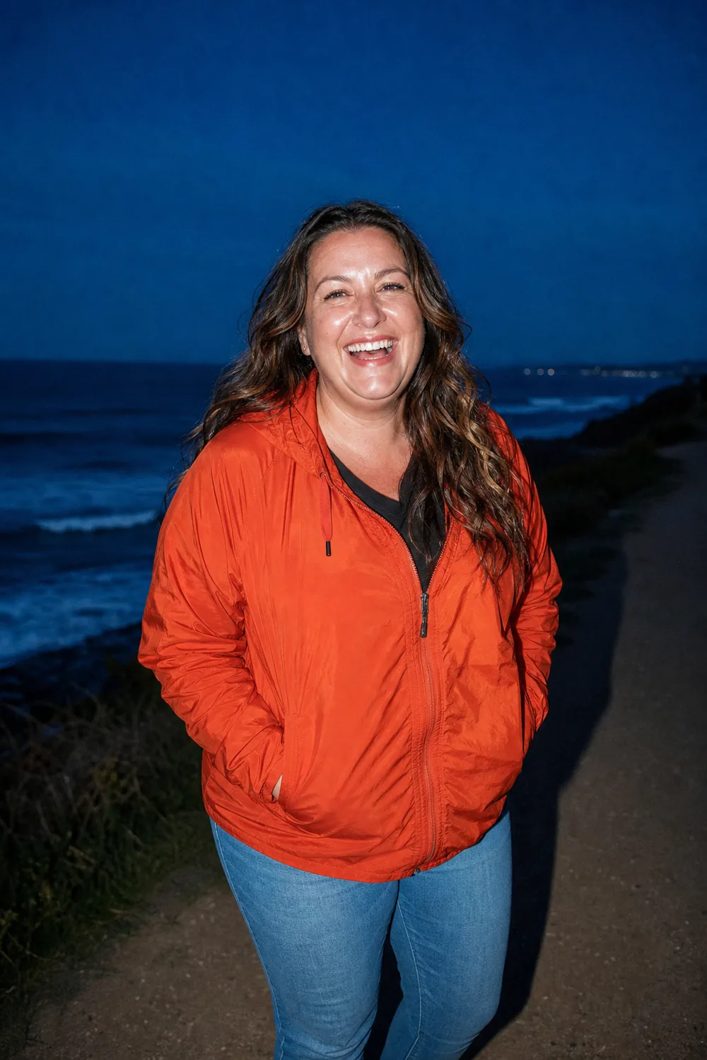 A woman in her forties laughing on a coastal path at dusk