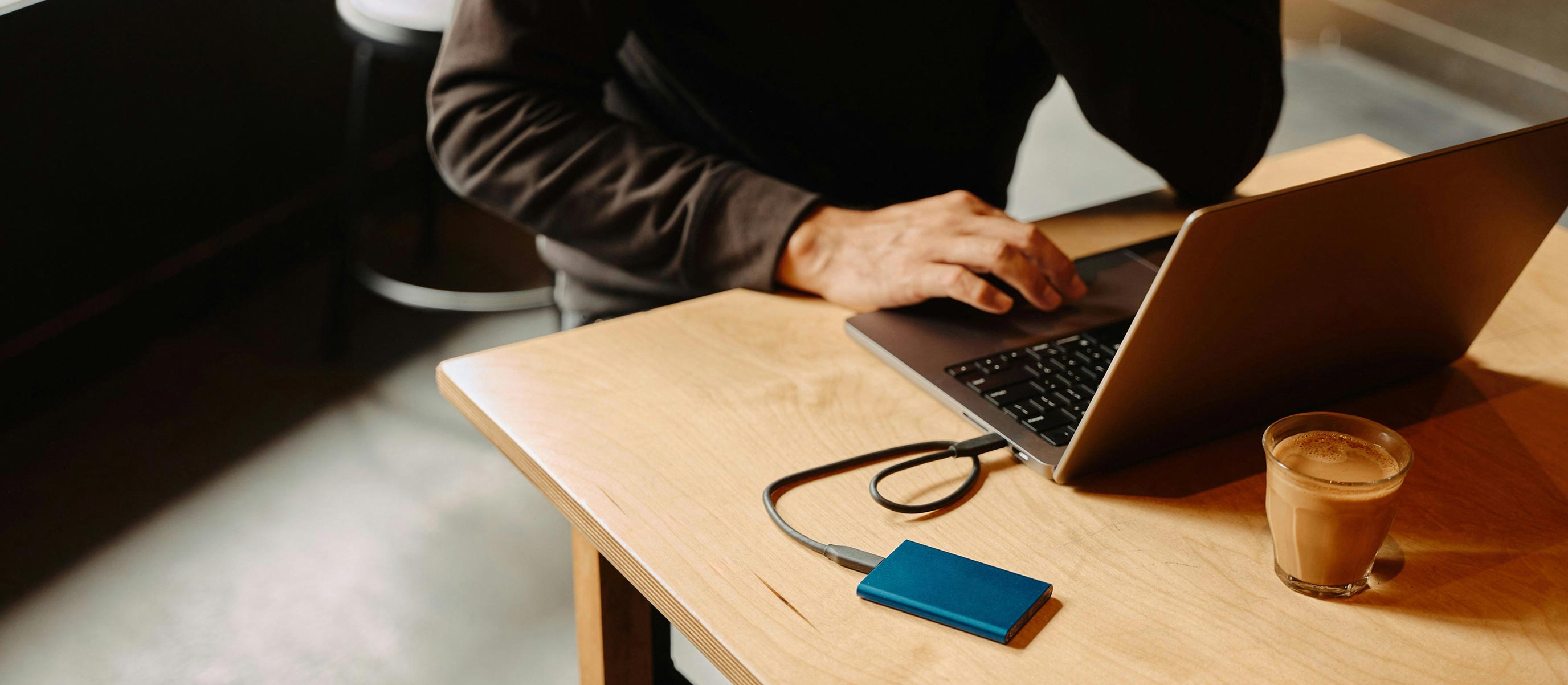 Person typing on a laptop connected to a blue Samsung external SSD on a wooden table with a glass of coffee nearby.
