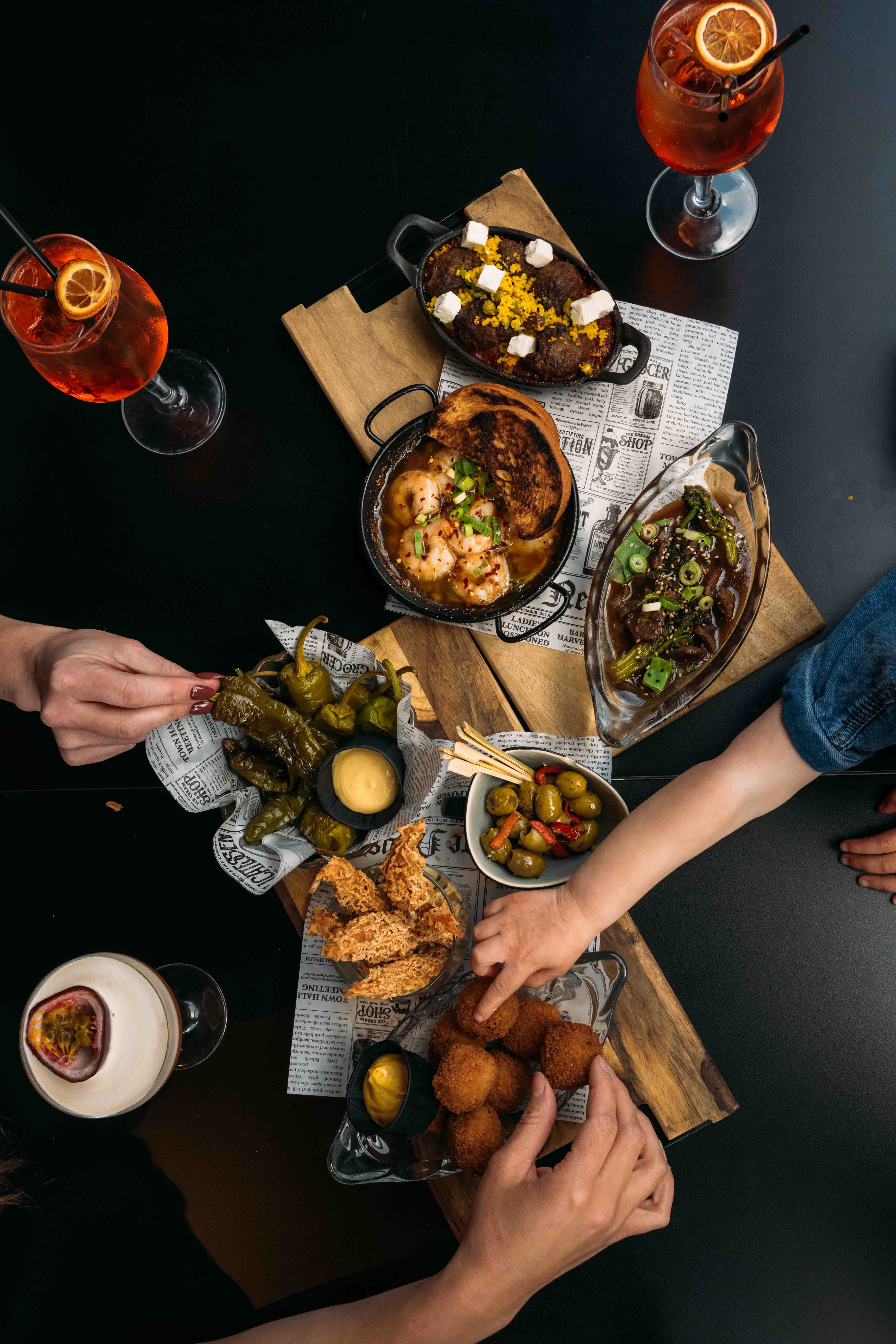 Bovenaanzicht van een tafel vol deelgerechtjes waar meerdere handen tegelijk uit pakken, met spritz-cocktails ernaast
