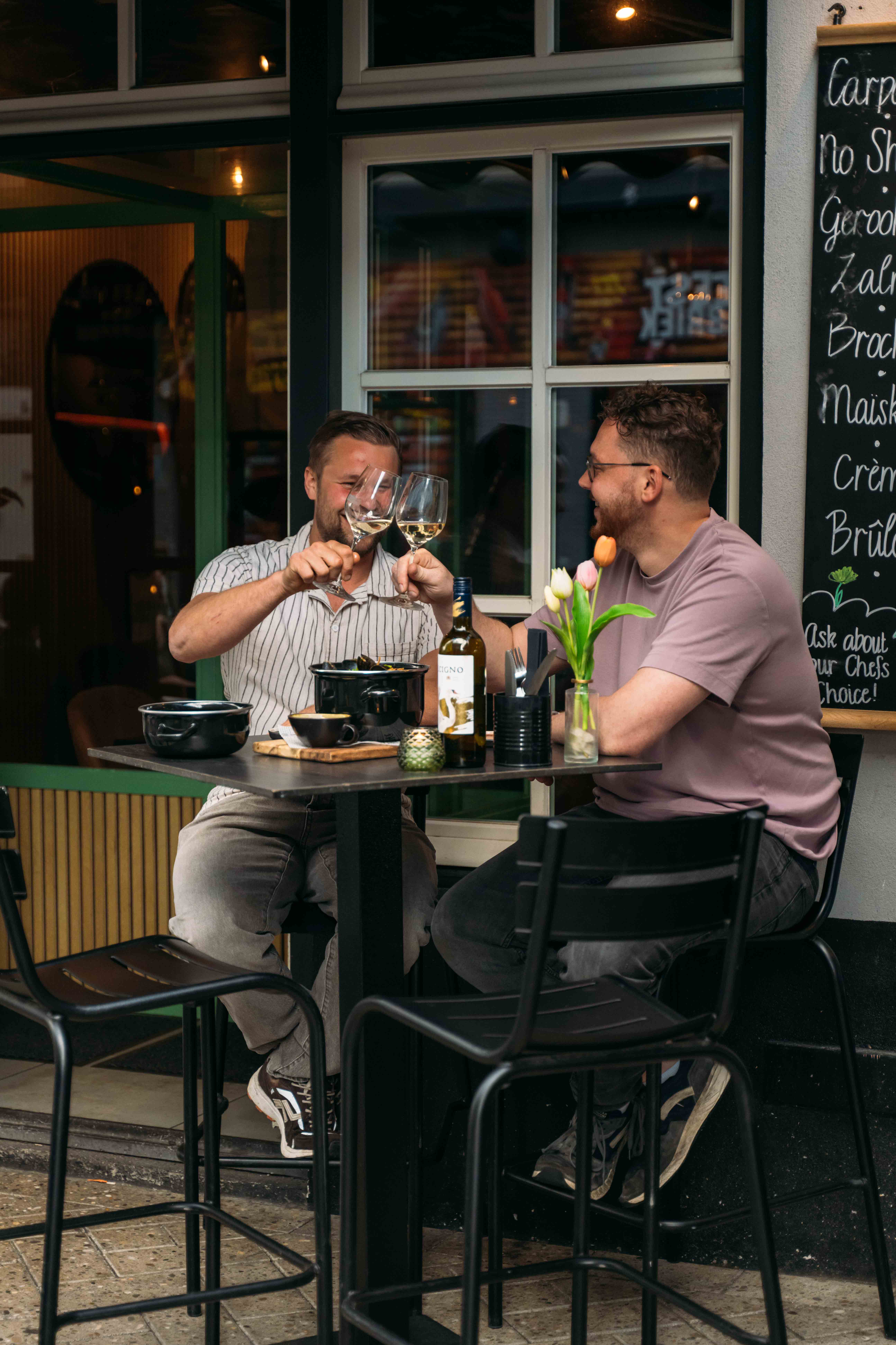 Twee gasten proosten met een glas witte wijn aan een tafeltje op het terras voor de zaak