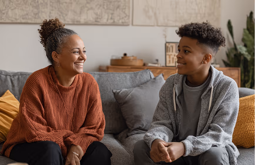 Smiling woman and teenage boy sitting and talking on a gray couch in a cozy living room.