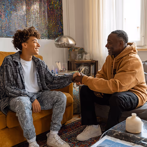 Two men smiling and shaking hands while sitting on couches in a cozy living room.