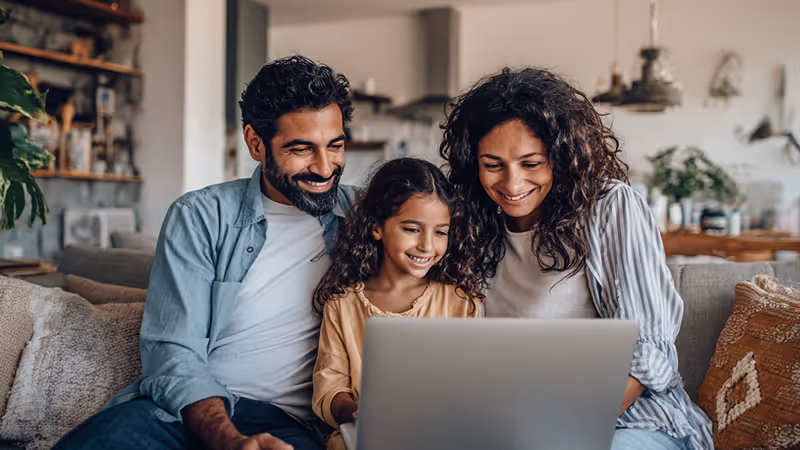 Smiling family of three sitting on a couch looking at a laptop screen together.