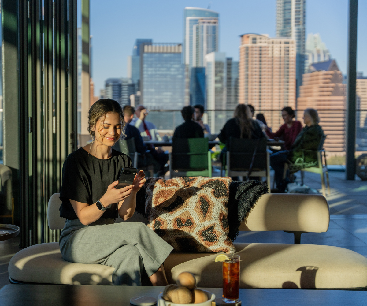 A woman sits on a couch in a high rise office building look at her smartphone.