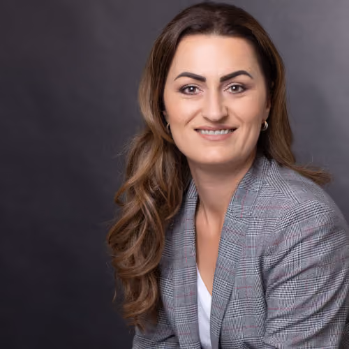 Portrait einer lächelnden Frau mit braunen Haaren und kariertem Blazer vor dunklem Hintergrund.