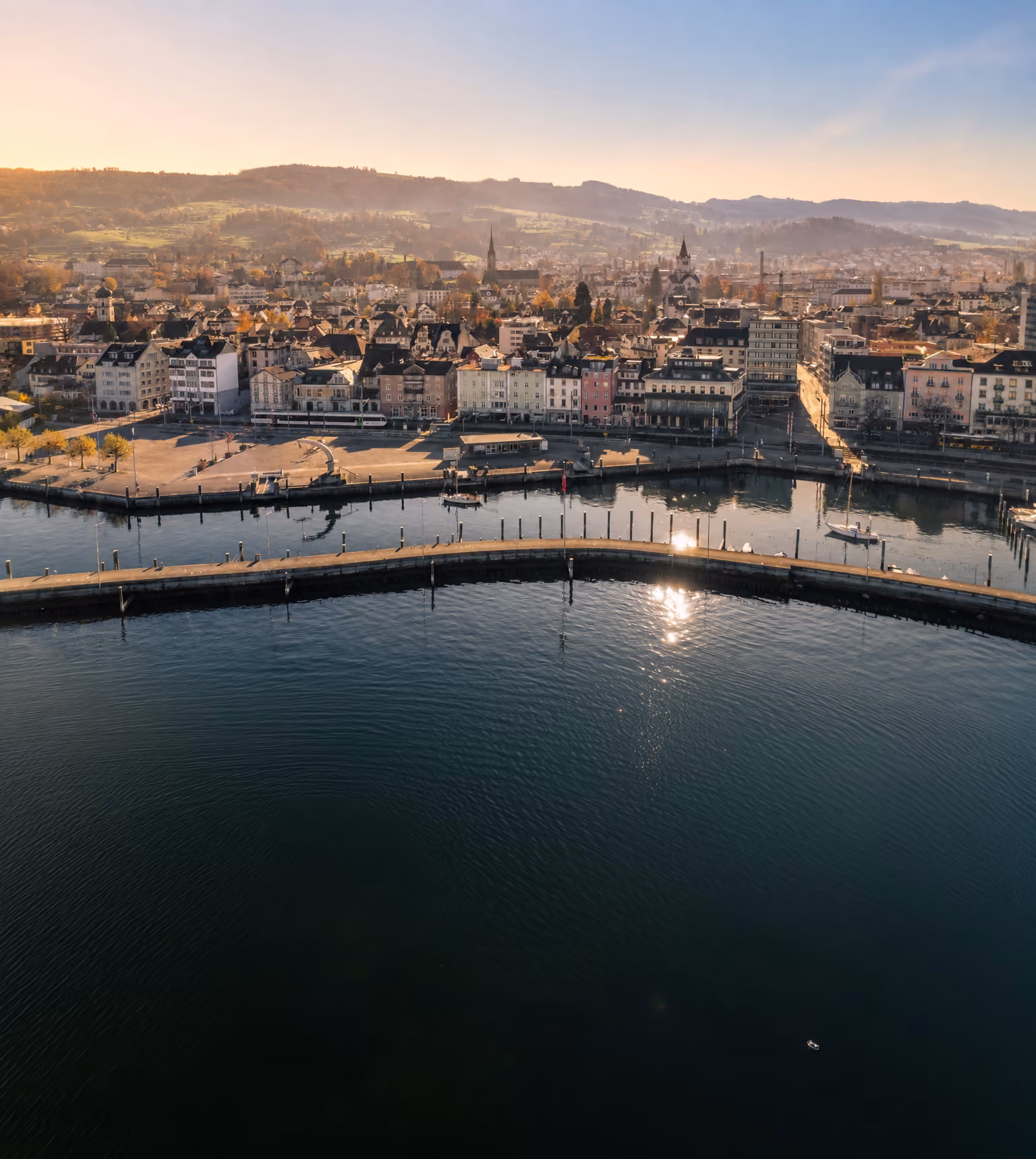Stadt am Ufer mit Bootsanleger und Hügeln im Hintergrund bei Sonnenuntergang.