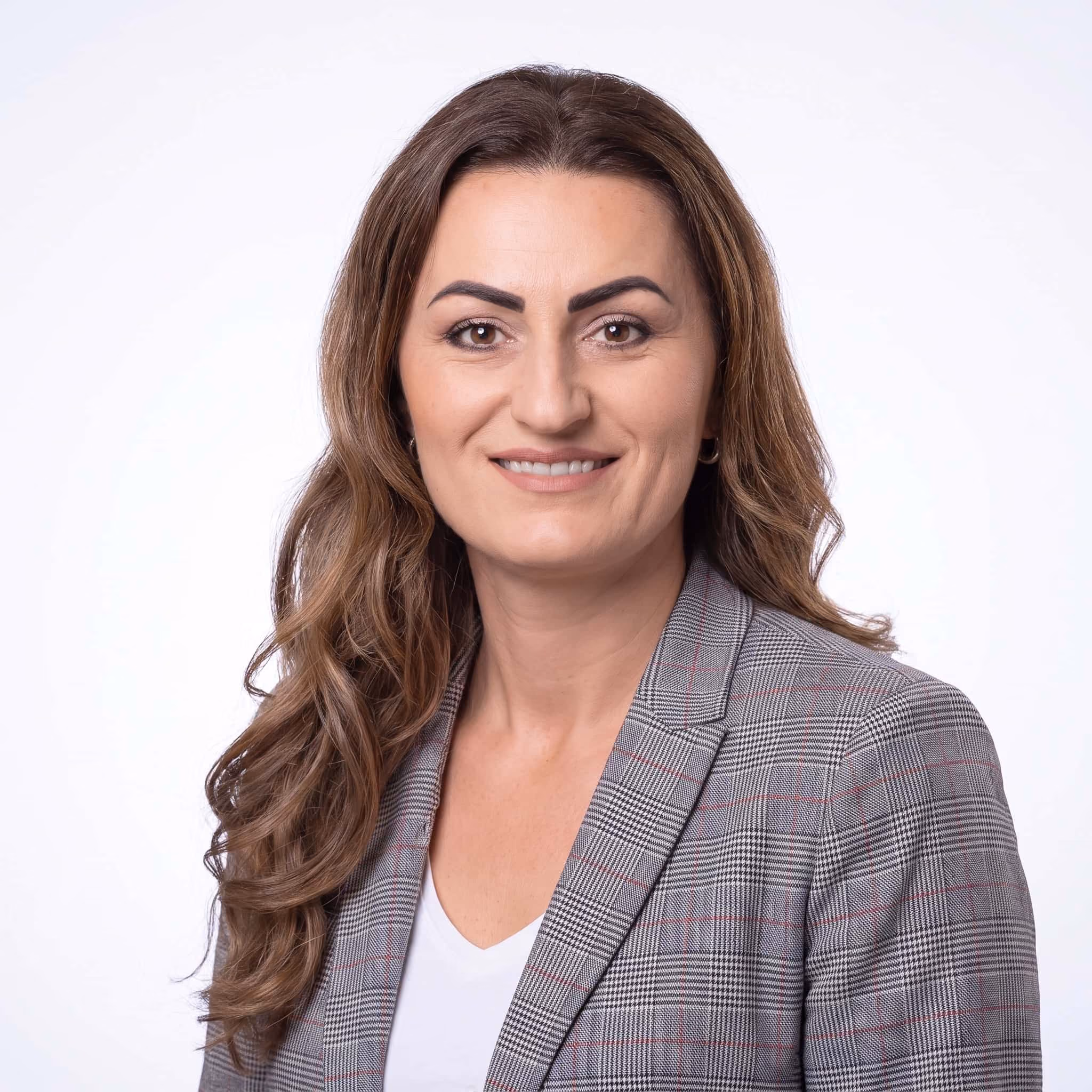 Porträt einer lächelnden Frau mit langen braunen Haaren, kariertem Blazer und weissem Top vor weissem Hintergrund.