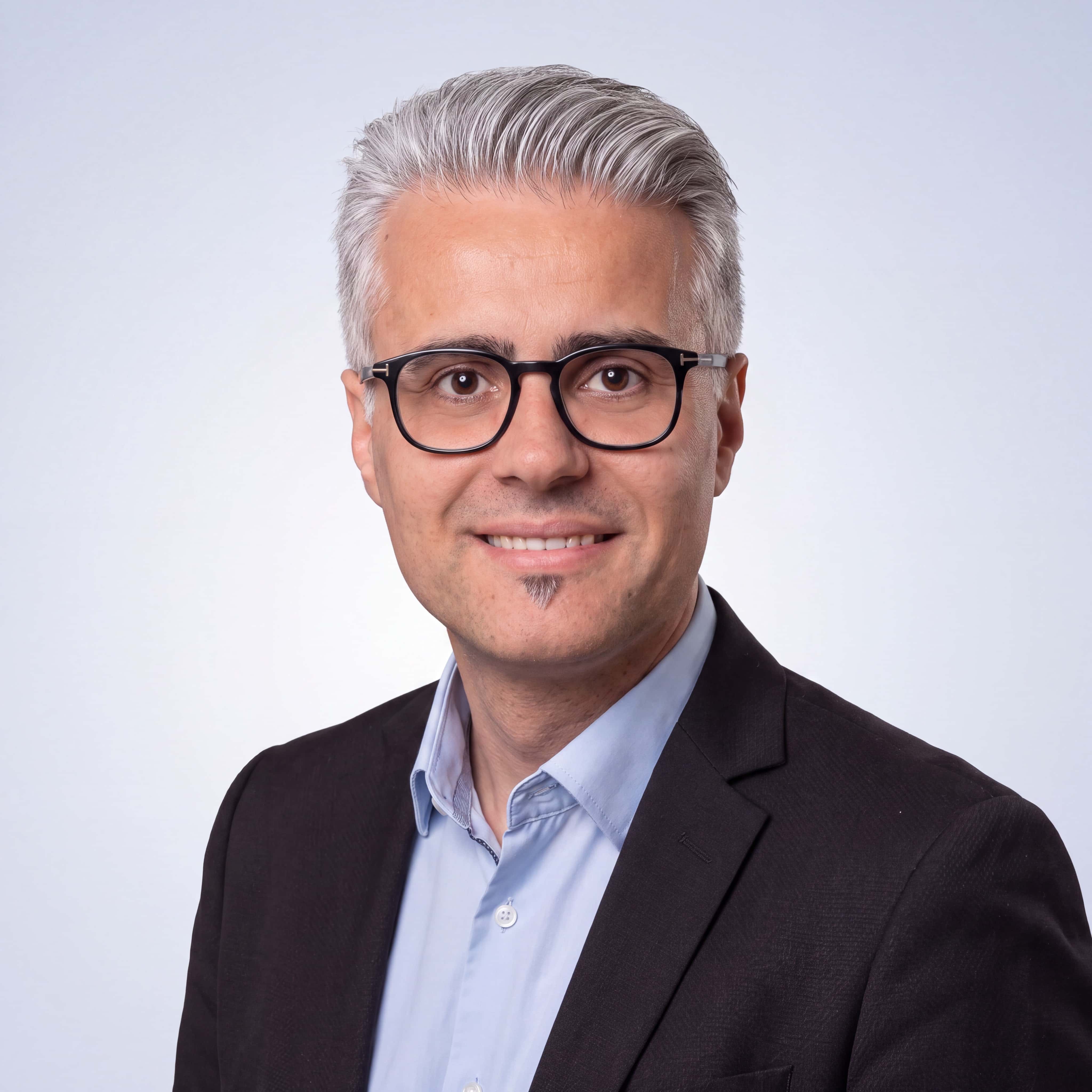 Mann mit grauen Haaren, Brille, blauem Hemd und dunklem Sakko lächelt vor hellgrauem Hintergrund.