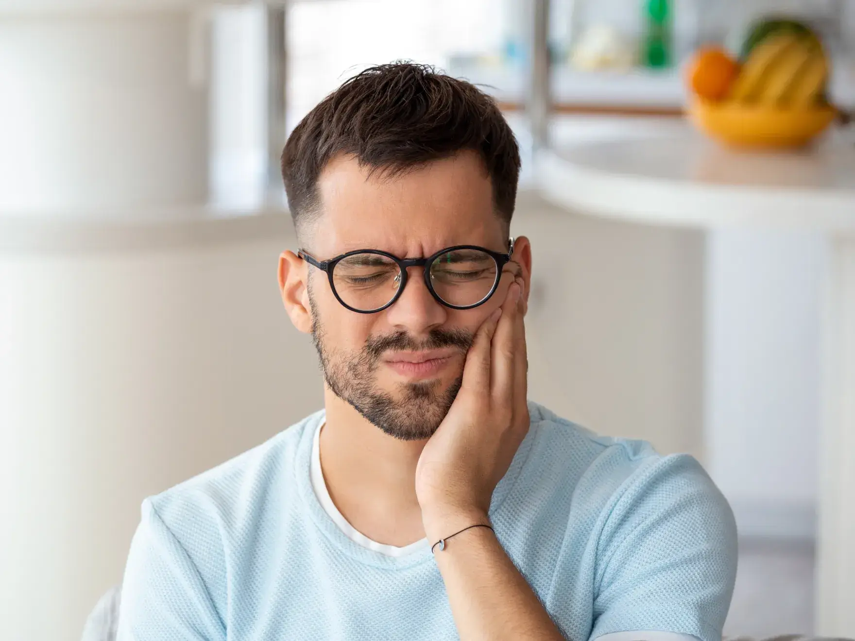 A photo of a man in glasses, grimacing, and holding his cheek with his hand.