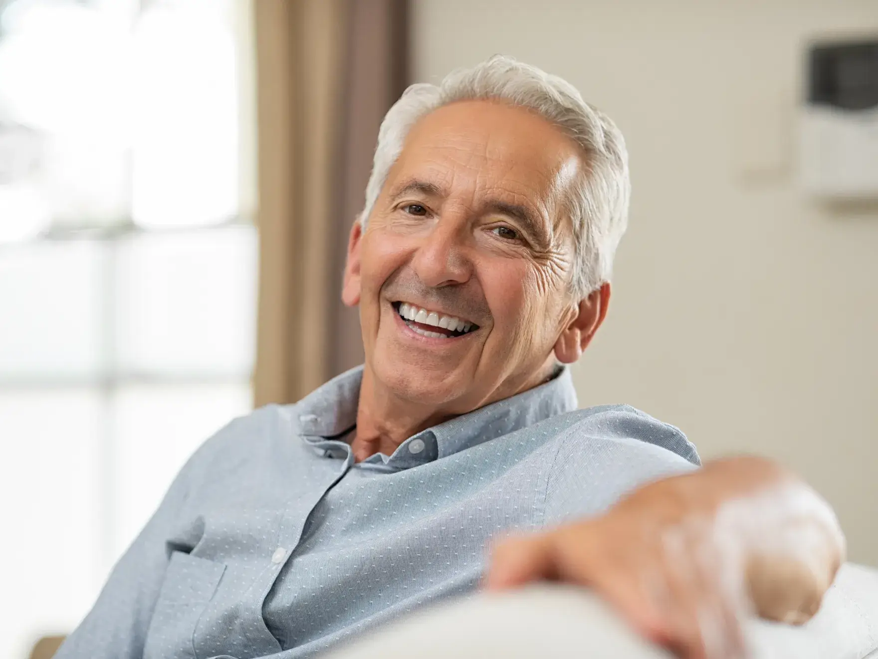 A photo of an elderly man with gray hair smiling, wearing a light blue shirt, and sitting comfortably indoors.