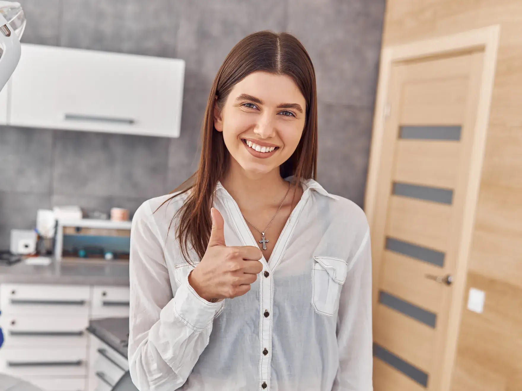 Photo of a smiling woman indoors, wearing a white shirt, giving a thumbs-up with a blurred kitchen in the background.