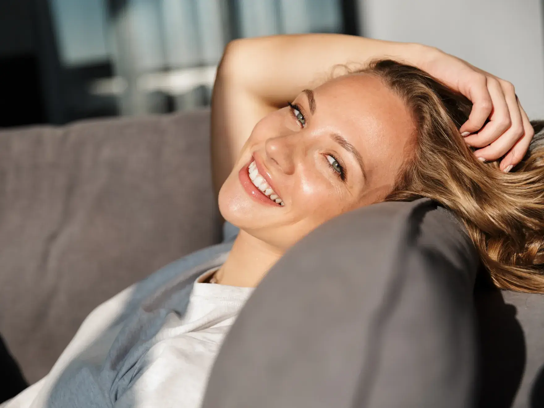 A photo of a smiling person with light hair, lounging on a couch, sunlight on their face.