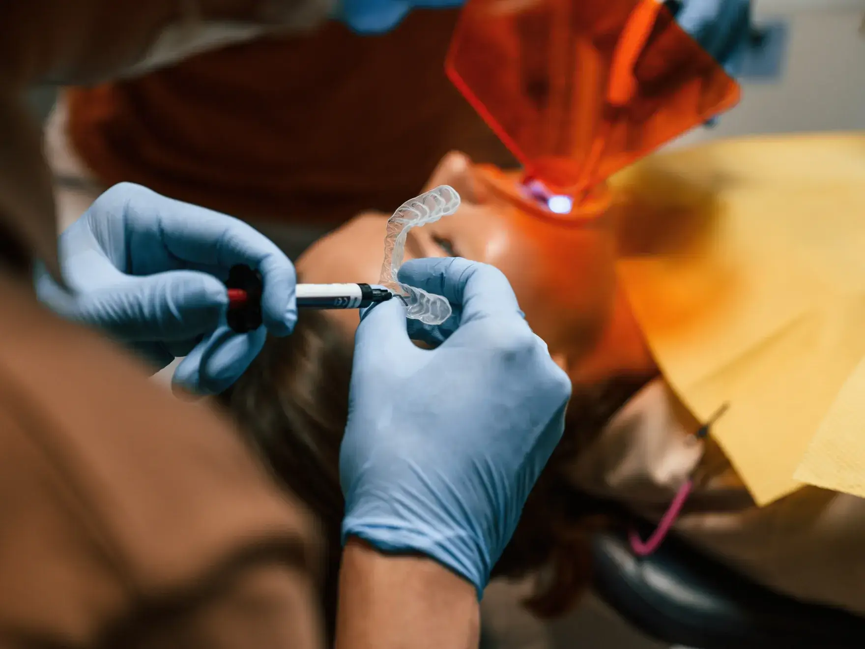 Dentist making dental impression on patient; blue gloves, orange shield; clinical setting.