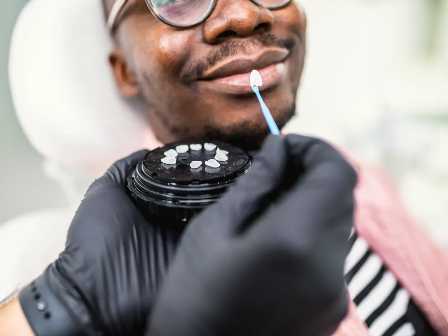 A close-up photo of a smiling person in glasses, with someone holding dental tools near their mouth.