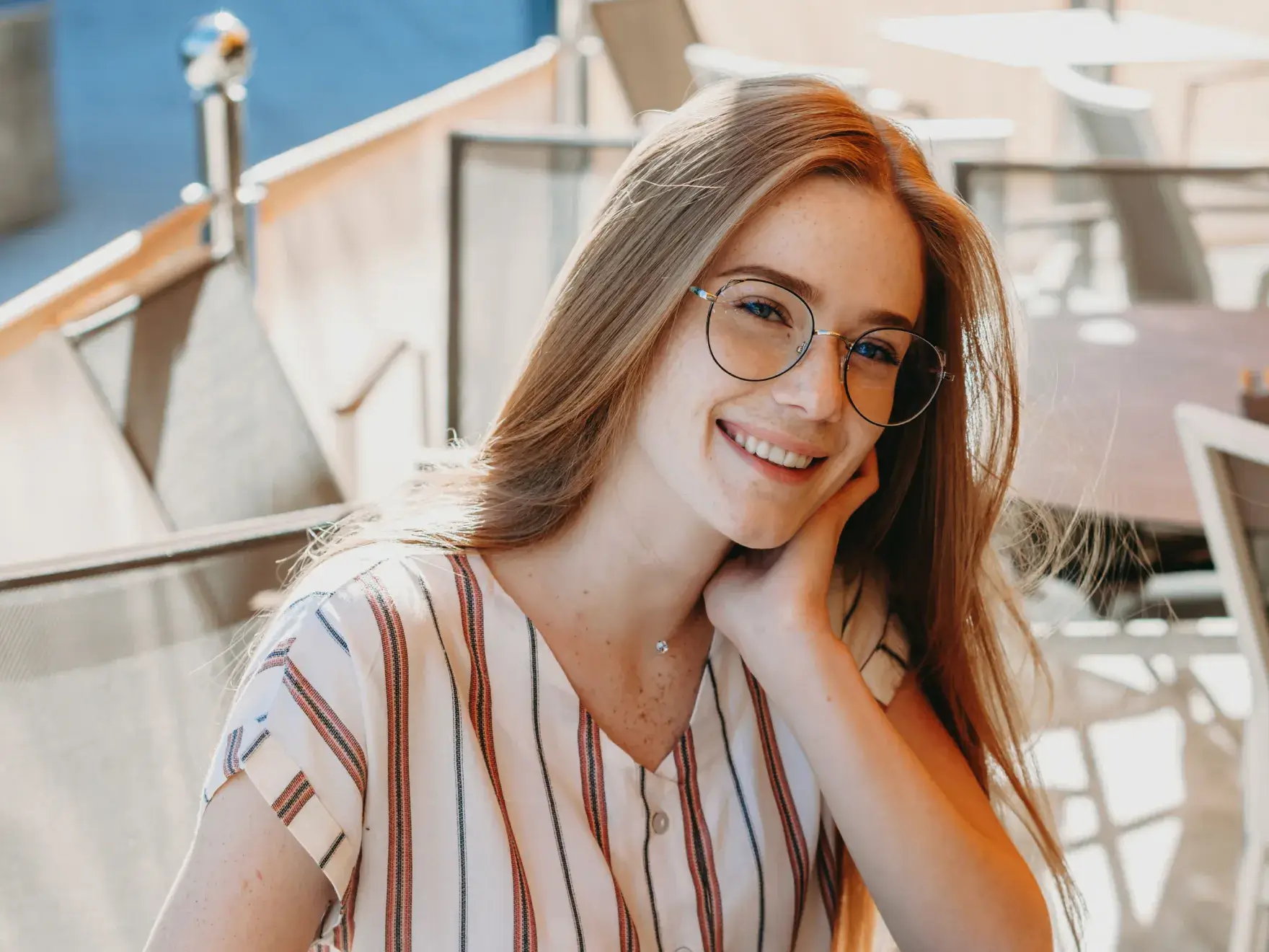 A photo of a smiling woman with long hair, wearing glasses and a striped shirt, sitting outdoors.