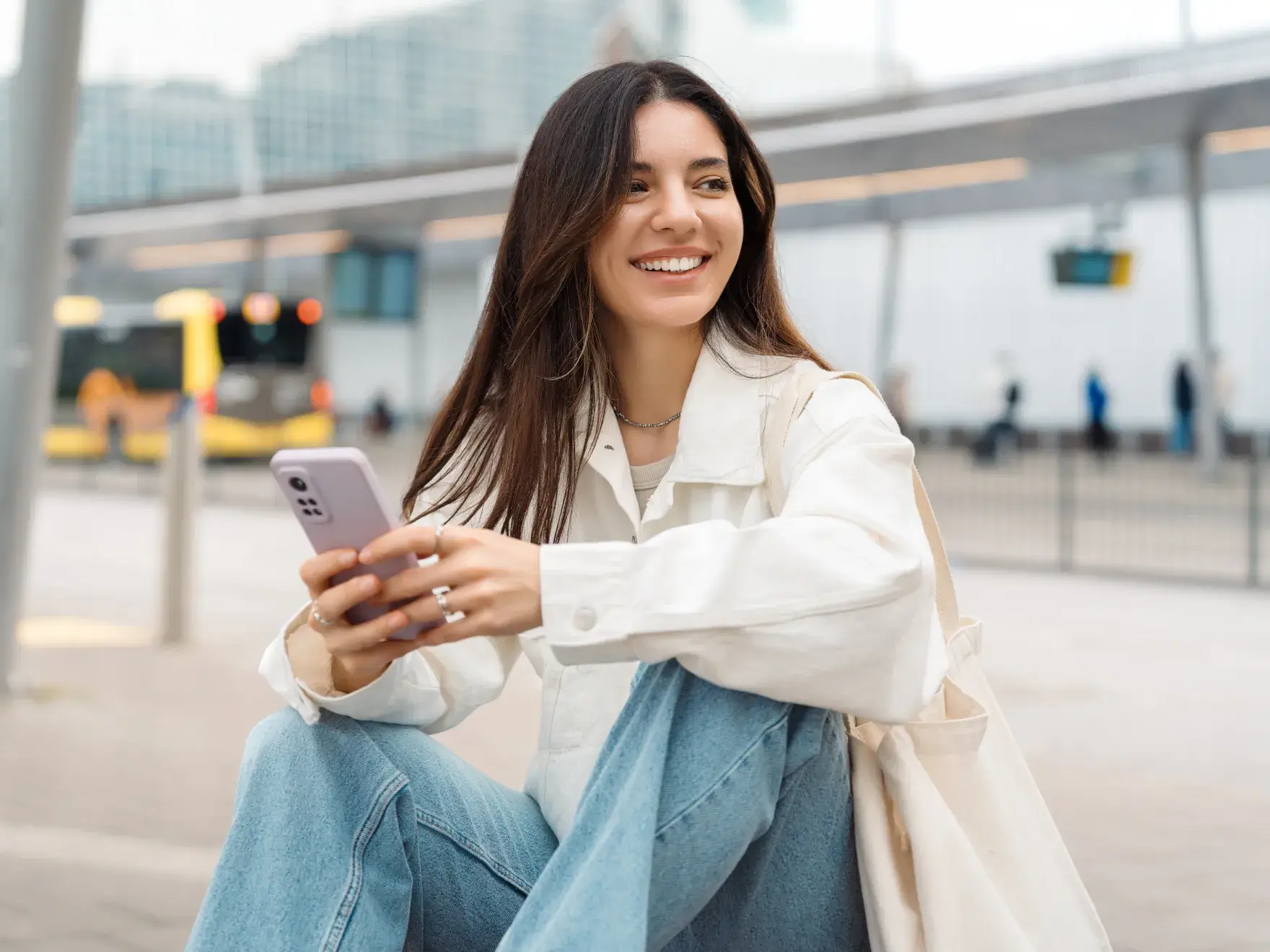A smiling woman in a white jacket and jeans sits outdoors with a smartphone.