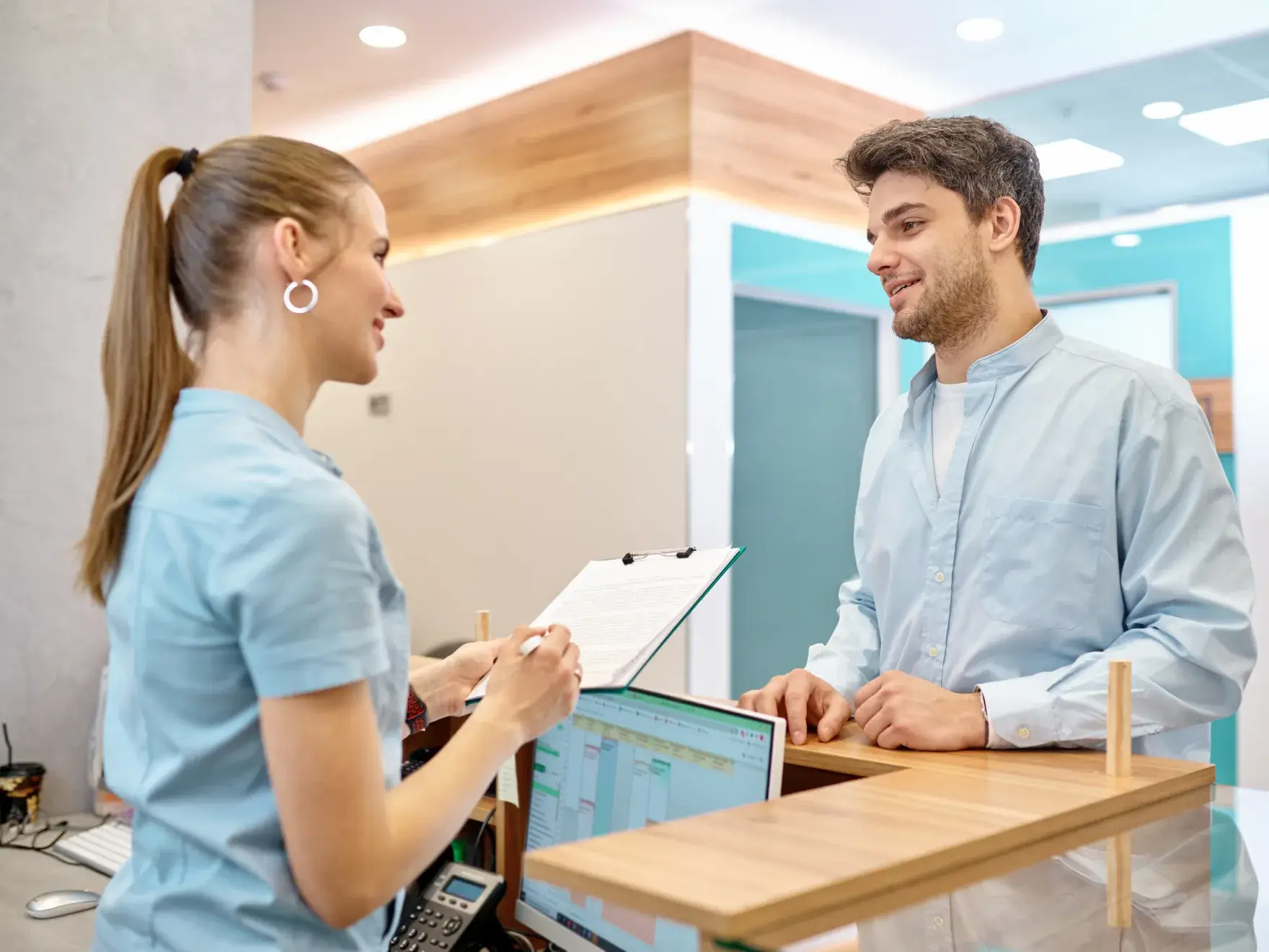 A photo of a woman and a man in a dental office, both in light blue uniforms, near a reception desk.